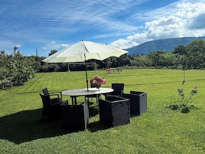 Een terras met een tafel en stoelen onder een grote parasol, omringd door een verzorgd gazon en bomen.