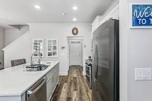 A modern kitchen with a stainless steel refrigerator, a built-in oven, and a sink with a dish rack. There is a staircase leading to another level, and a welcome sign on the wall. - The Reverie - Downtown Pensacola - Convenient Corner Lot - Great For Business Travelers - Fast WiFi (Pensacola)