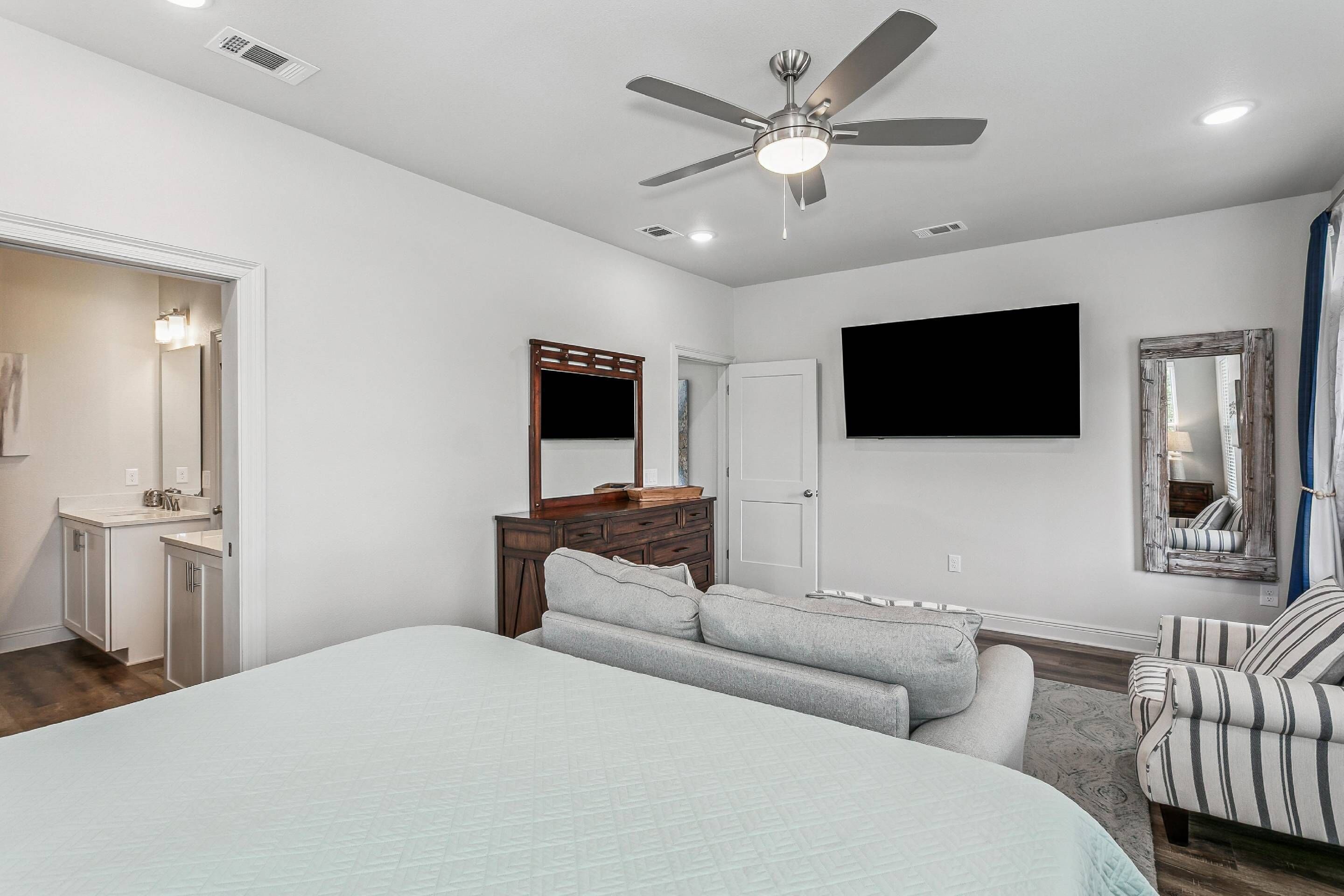A bedroom with a bed, a dresser, a mirror, a ceiling fan, and a TV mounted on the wall.