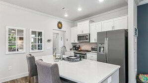 A modern kitchen with white cabinetry, a central island with bar stools, a stainless steel refrigerator, and a microwave. - The Reverie - Downtown Pensacola - Convenient Corner Lot - Great For Business Travelers - Fast WiFi (Pensacola)