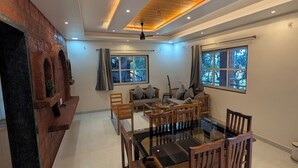A modern dining area with a glass-top table and wooden chairs, a ceiling fan, and a view of the outdoors through large windows.