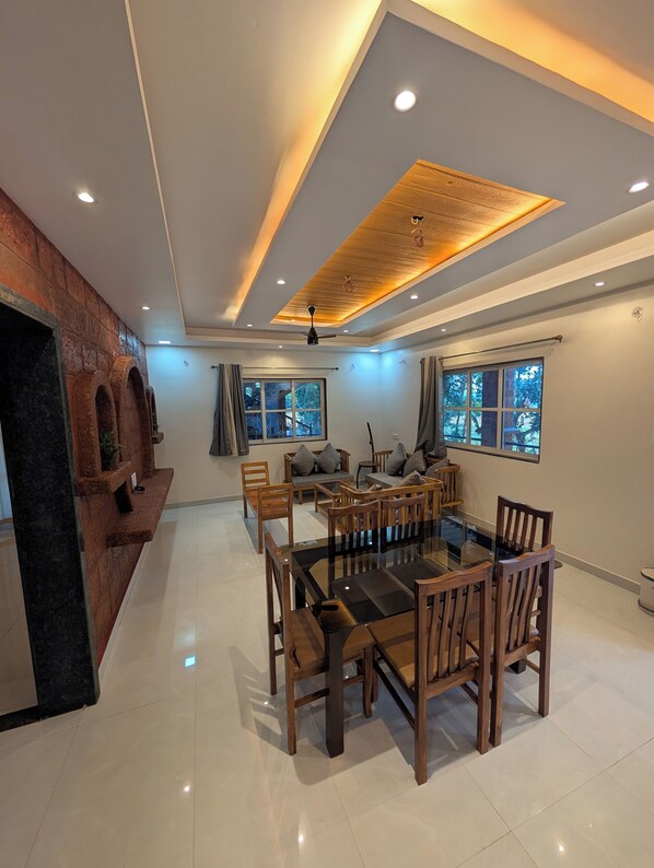 A modern dining area with a glass-top table and wooden chairs, a ceiling fan, and a view of the outdoors through large windows.