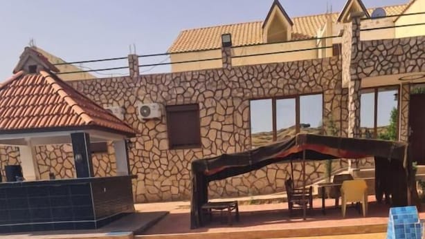 A swimming pool with clear blue water, a stone building with a tiled roof, and a covered outdoor seating area.