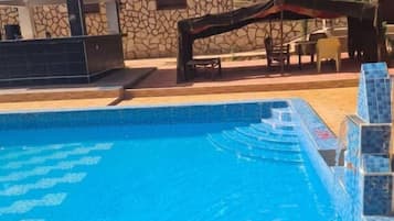 Une piscine à l’eau cristalline, un bâtiment en pierre avec un toit en tuiles et un espace extérieur aménagé avec des sièges, protégé par un auvent.