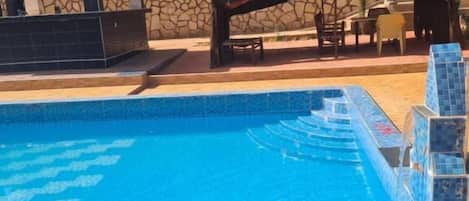 A swimming pool with clear blue water, a stone building with a tiled roof, and a covered outdoor seating area.