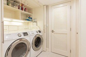 Un espace buanderie équipé d’un lave-linge et d’un sèche-linge Samsung, d’une étagère contenant des produits de nettoyage et d’une porte fermée avec des stores.