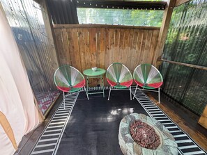 A covered patio with a wooden fence, three green and white wire chairs, a small round table, and a fire pit with rocks.