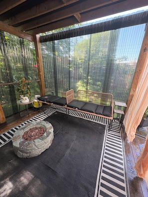 A covered outdoor seating area with a stone fire pit, cushioned seating, and a view through a bamboo screen.