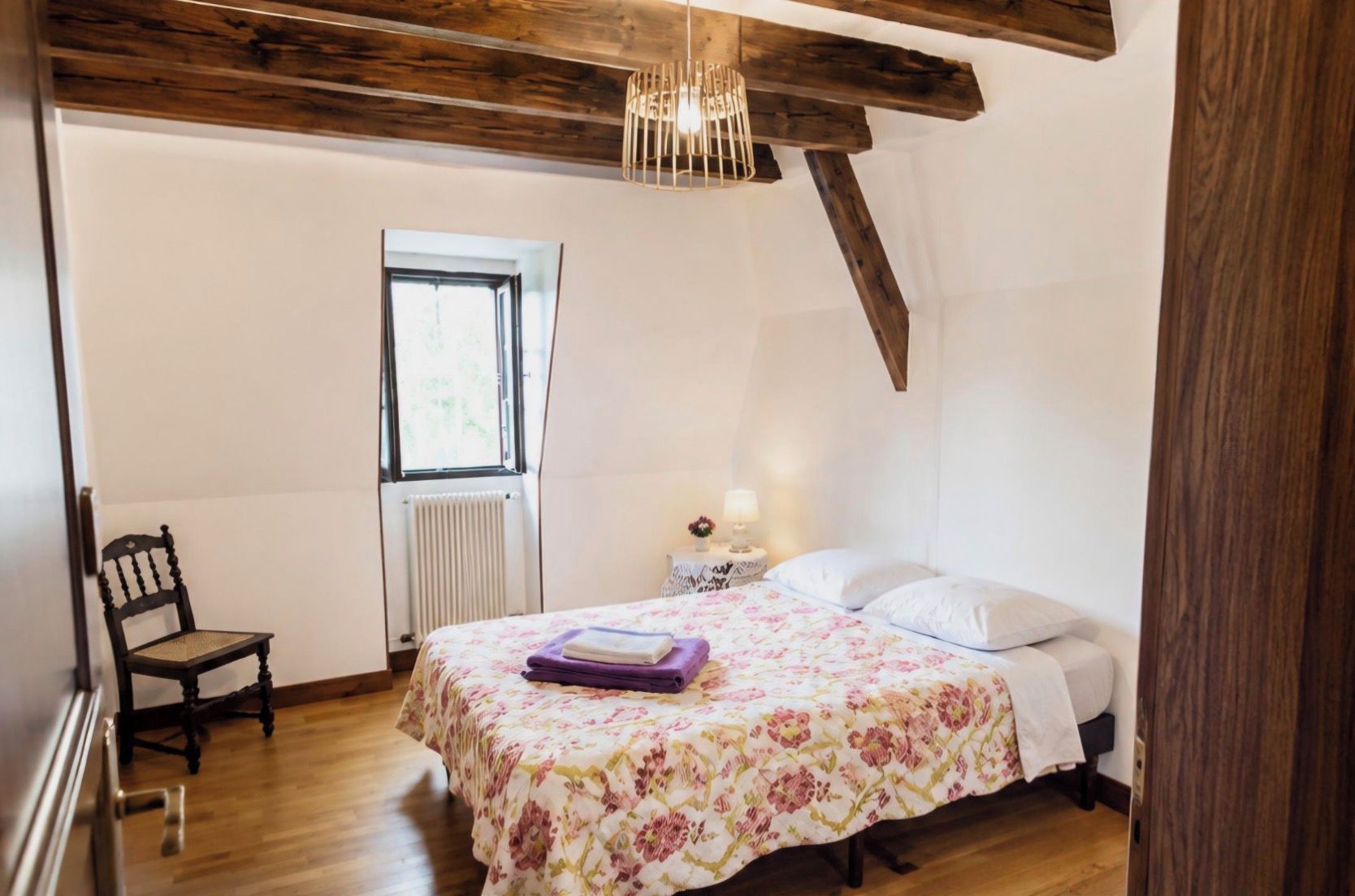 A bedroom with a wooden ceiling, a bed with floral bedding, a chair, a bedside table with a lamp, and a window.