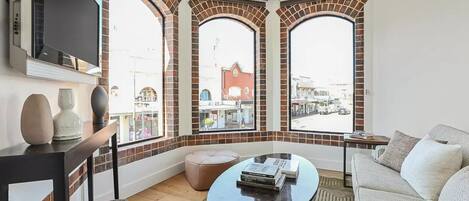 A modern living room with a large TV, a round coffee table, and a sofa. There are three arched windows with brick detailing, and a view of the street outside.