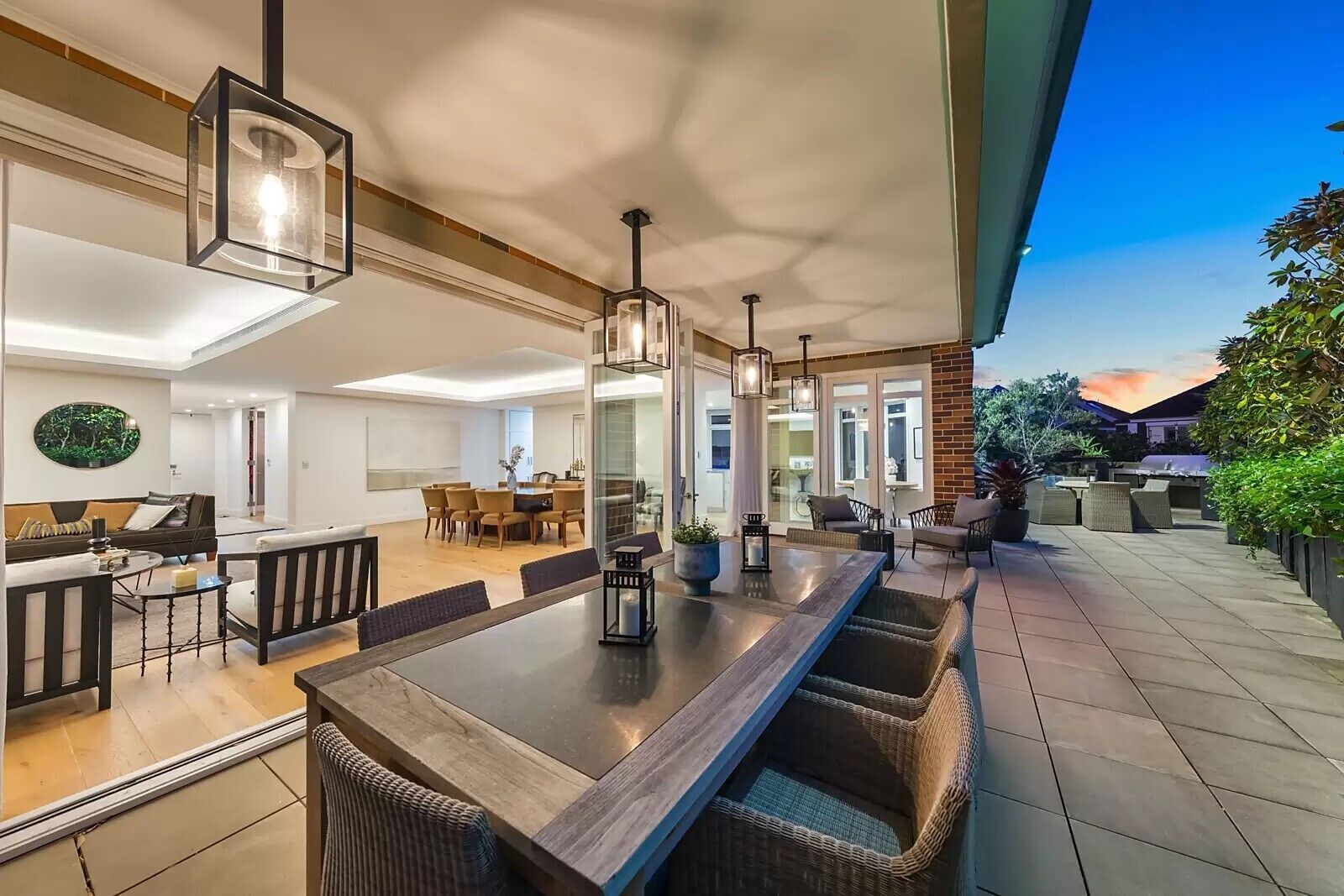 A modern outdoor dining area with a long table, wicker chairs, and pendant lighting.