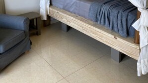 En säng för en person med en träram och ett sänghimmel, ett litet sängbord och en grå soffa i ett rum med kakelgolv.
