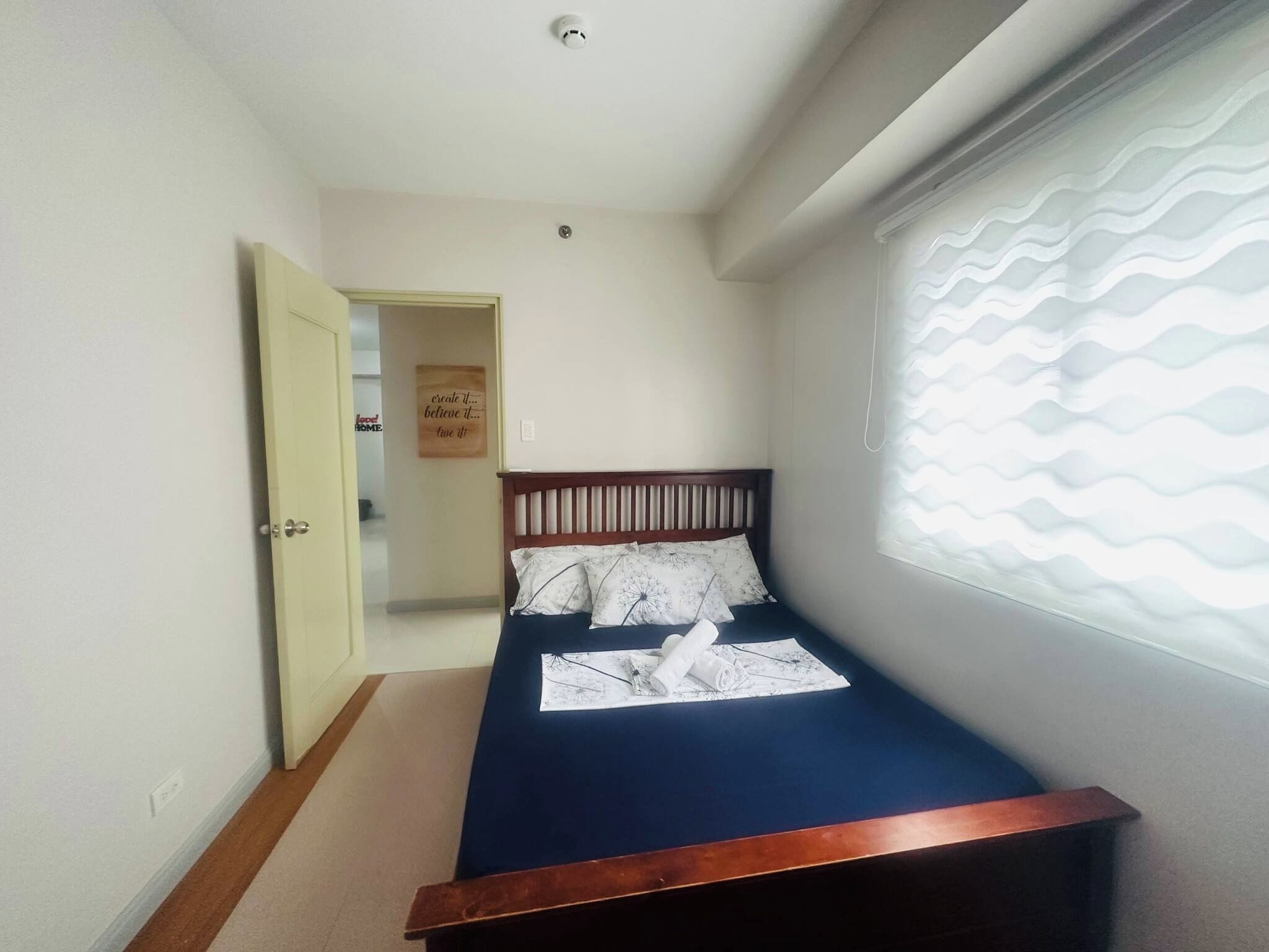 A narrow room with a single bed, a door leading to another room, and a window with a wavy pattern blind.