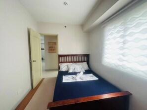 A narrow room with a single bed, a door leading to another room, and a window with a wavy pattern blind.