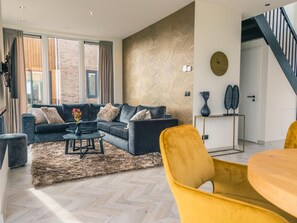 Ein modernes Wohnzimmer mit einem Ecksofa, einem Couchtisch und einem gelben Sessel. Eine Treppe befindet sich rechts und ein großes Fenster mit Vorhängen ist vorhanden.