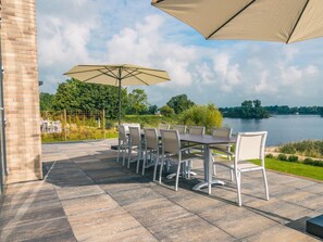 Eine Terrasse im Freien mit Tisch und Stühlen, einem großen Sonnenschirm und Blick auf einen See.