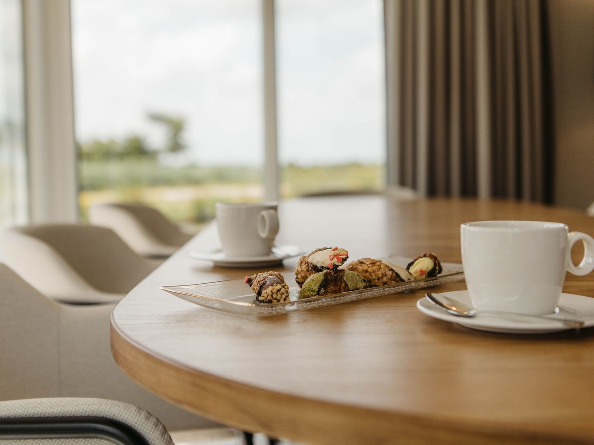 Una mesa con una bandeja de pasteles y dos tazas blancas, probablemente para café o té, en un interior moderno con vistas al exterior.
