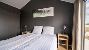 A modern bedroom with a large bed, two bedside tables with lamps, a dark wall, and a window with curtains. - Vakantiepark Aan de Maas - Maas Lande | 4p by Interhome (Kerkdriel)