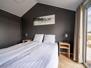 A modern bedroom with a large bed, two bedside tables with lamps, a dark wall, and a window with curtains. - Vakantiepark Aan de Maas - Maas Lande | 4p by Interhome (Kerkdriel)