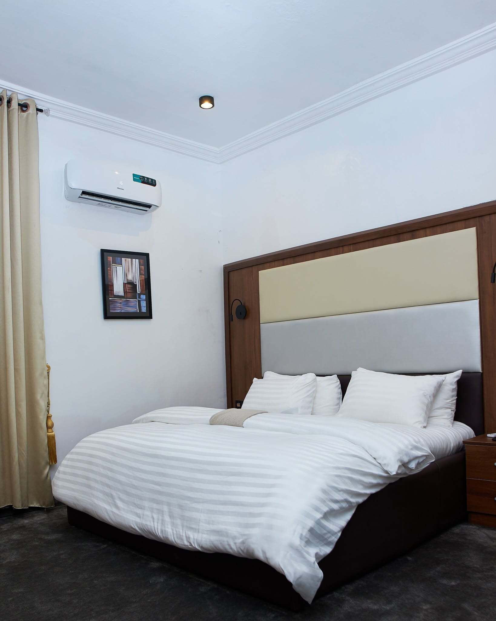 A bedroom with a bed, a picture on the wall, an air conditioning unit, and a ceiling light.