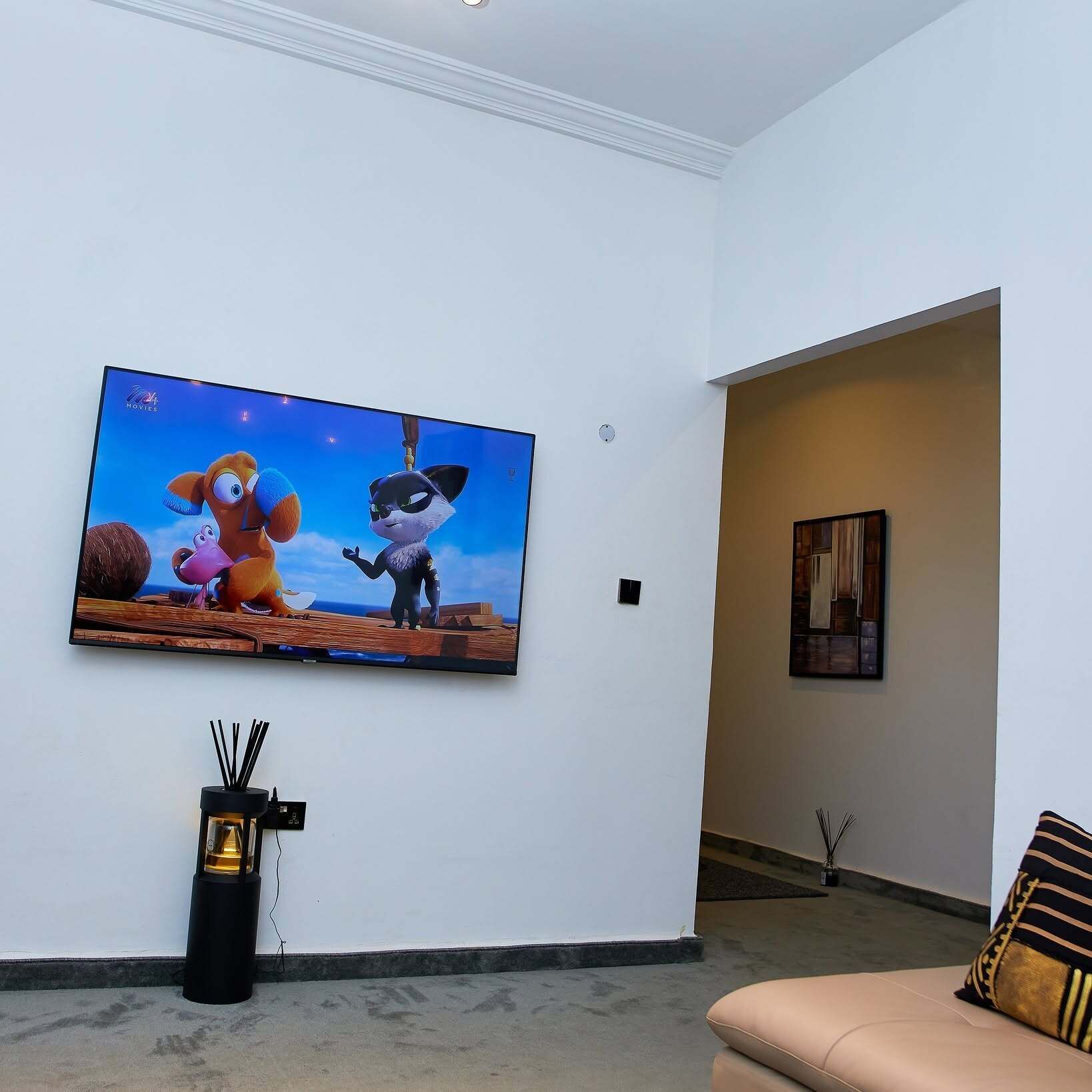 Una sala de estar con un televisor de pantalla plana grande montado en la pared, un altavoz negro y un sofá beige con un cojín a rayas.