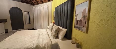 Un dormitorio con cama, mesita de noche, cabecero, ventana con cortinas e imagen colgada en la pared.