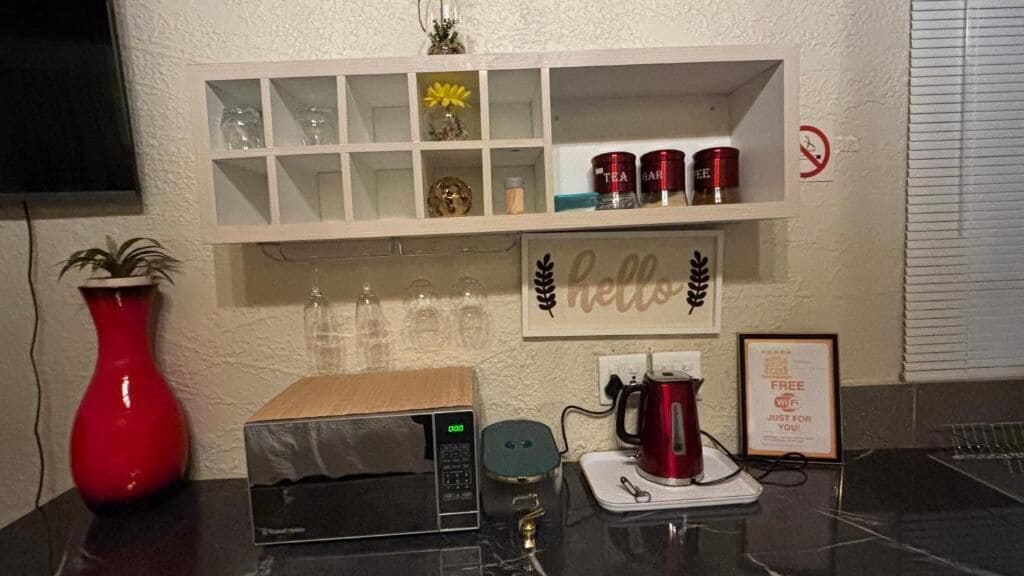 A kitchen area with a microwave, a red vase, a coffee maker, and a shelf with various items including jars and a 'hello' sign.