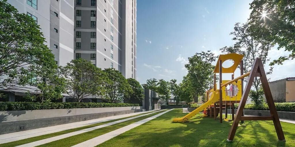 Un terrain de jeux avec un toboggan jaune et des balançoires, entouré d’une pelouse bien entretenue et d’arbres, avec un grand immeuble en arrière-plan.
