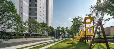 Un parque infantil con un tobogán amarillo y columpios, rodeado de césped cuidado y árboles, con un edificio alto al fondo.