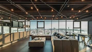 A modern lounge area with a large sofa, a coffee table, and a dining table with chairs. There is a large window offering a view of a cityscape.