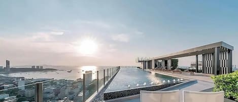 A rooftop pool with lounge chairs, overlooking a cityscape and a body of water.