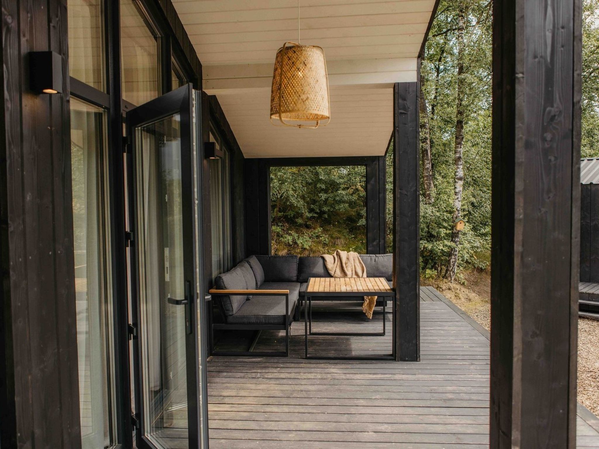 Een modern terras met een zithoek, inclusief een bank en een tafel, een hanglamp en uitzicht op het omliggende bos.