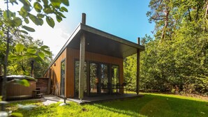 A modern wooden cabin with a glass enclosure, a patio, and a hot tub in a lush green setting. - De Wiltzangh - Bosrand Wellness Cabin | 2p by Interhome (Ruinen)