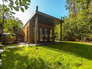A modern wooden cabin with a glass enclosure, a patio, and a hot tub in a lush green setting. - De Wiltzangh - Bosrand Wellness Cabin | 2p by Interhome (Ruinen)