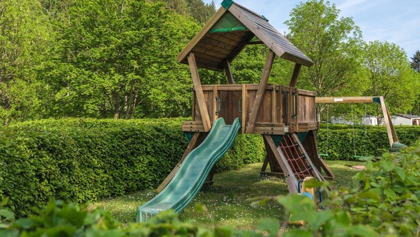 A wooden playhouse with a slide and climbing net, surrounded by greenery and a hedge.