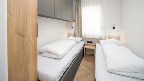 A hotel room with two single beds, a wooden wardrobe, a bedside table, and a window with sheer curtains.