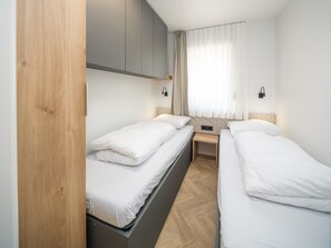 A hotel room with two single beds, a wooden wardrobe, a bedside table, and a window with sheer curtains.