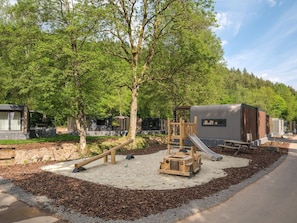A playground with a slide and climbing structure, surrounded by trees and a gravel area.