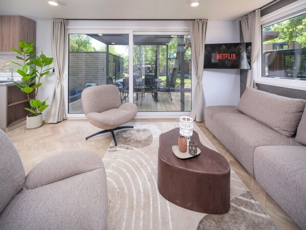 A modern living room with a sofa, armchair, and coffee table. There is a television mounted on the wall and a large window with curtains.