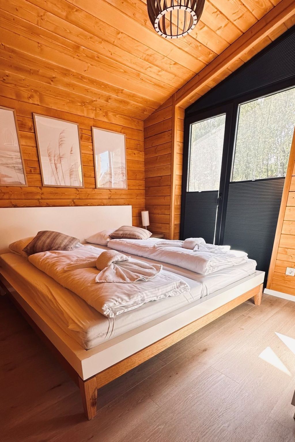 Een slaapkamer met houten lambrisering, een bed, afbeeldingen in lijsten en een raam met zonwering.