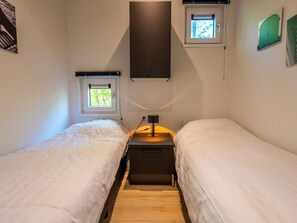 Ein kleines, helles Zimmer mit zwei Einzelbetten, einem Nachttisch mit Lampe und zwei Fenstern mit Jalousien.