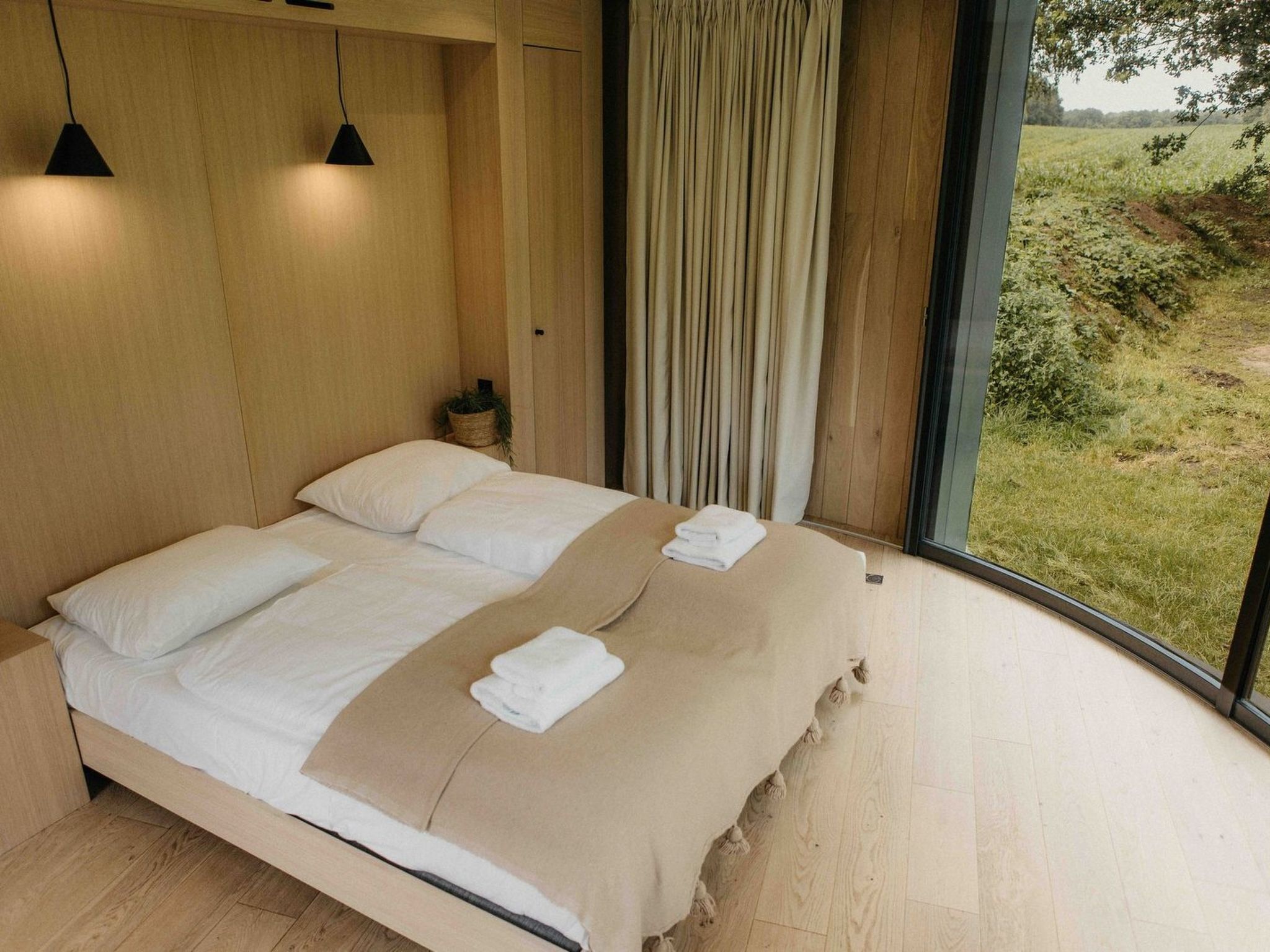 A modern bedroom with a large bed, two bedside lamps, a view of the outdoors, and a wooden floor.