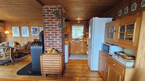 A cozy cabin interior with a brick fireplace, wooden furniture, a kitchen area with a refrigerator and microwave, and a dining area with wicker chairs.