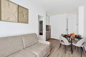 Ein modernes Wohnzimmer mit einem beigefarbenen Sofa, einem Essbereich mit Glastisch und Stühlen, und eine durch eine offene Tür sichtbare Küche.