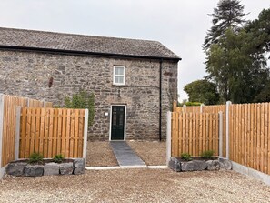 A stone building with a black door, wooden gates, and a gravel pathway leading to the entrance.