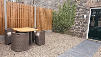 A patio with a wooden table and wicker chairs, a gravel path, a stone wall, and a green door.
