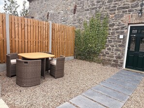 Un patio con una mesa de madera y sillas de mimbre, un sendero de grava, un muro de piedra y una puerta verde.