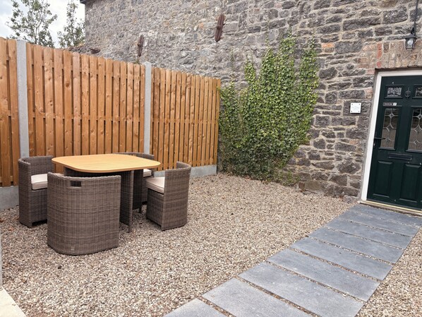A patio with a wooden table and wicker chairs, a gravel path, a stone wall, and a green door.