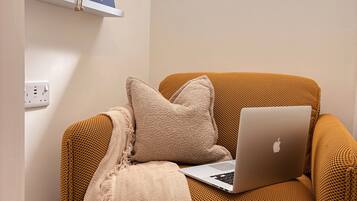 A cozy seating area with a laptop on a textured sofa, a decorative wall shelf with books and figurines, and a window with a view of the sky.