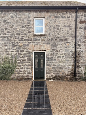 Un bâtiment en pierre avec une porte noire, une petite fenêtre au-dessus de la porte et un chemin de gravier menant à l’entrée.